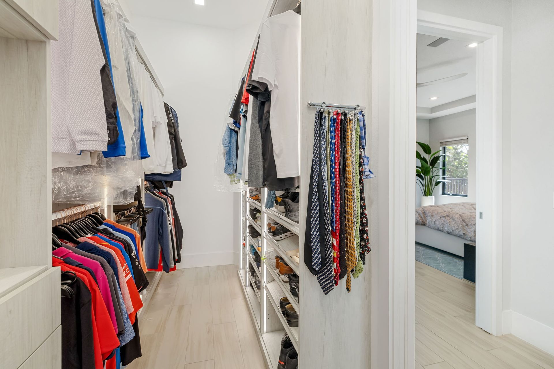 Bright walk-in closet with hanging clothes, shelves, and an open doorway to a bedroom