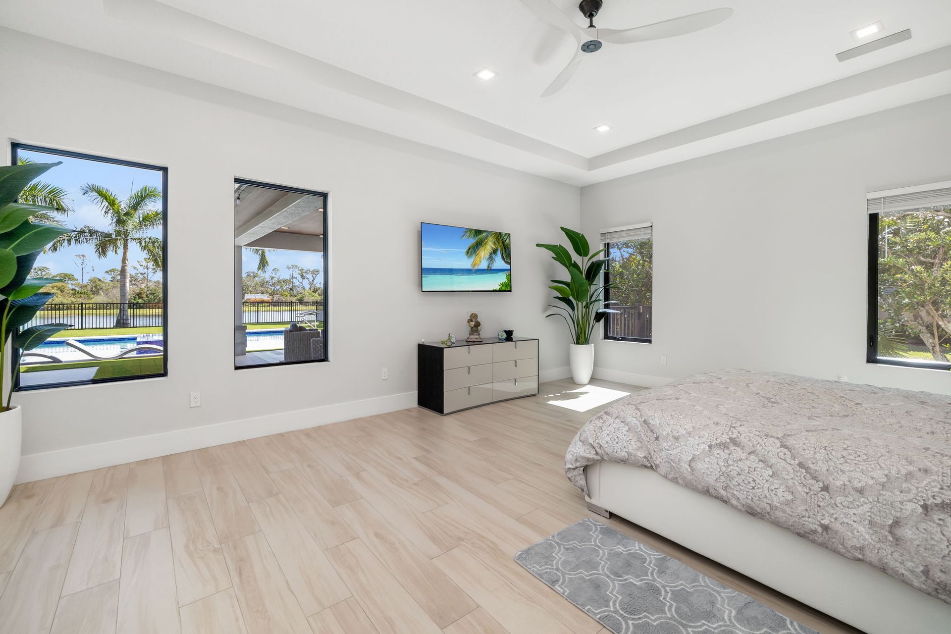 Bright modern bedroom with large windows, white walls, light wood floors, and a bed beside a TV and dresser.