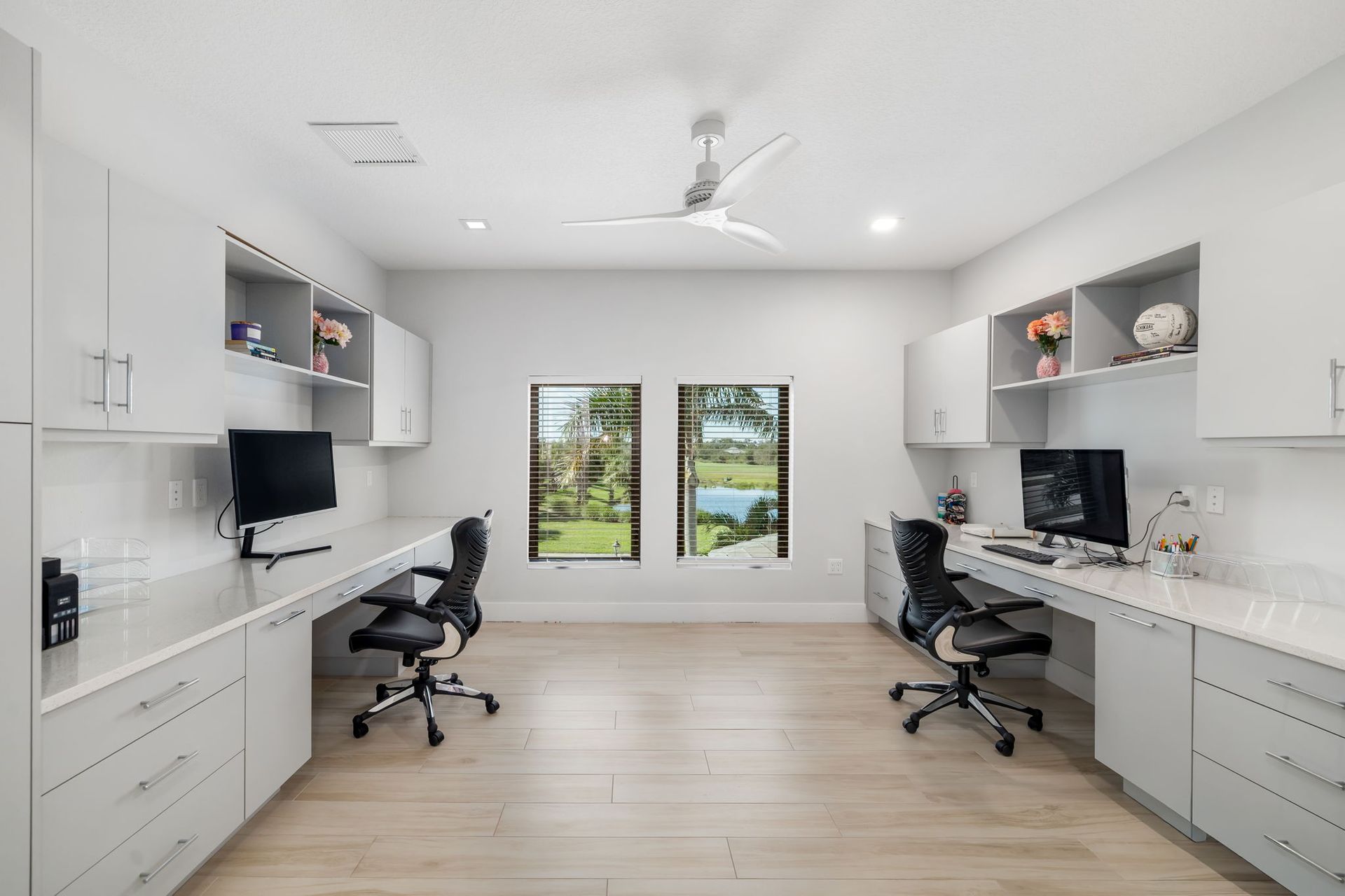 Bright modern dual-desk home office with white cabinets, black chairs, and garden view through French doors