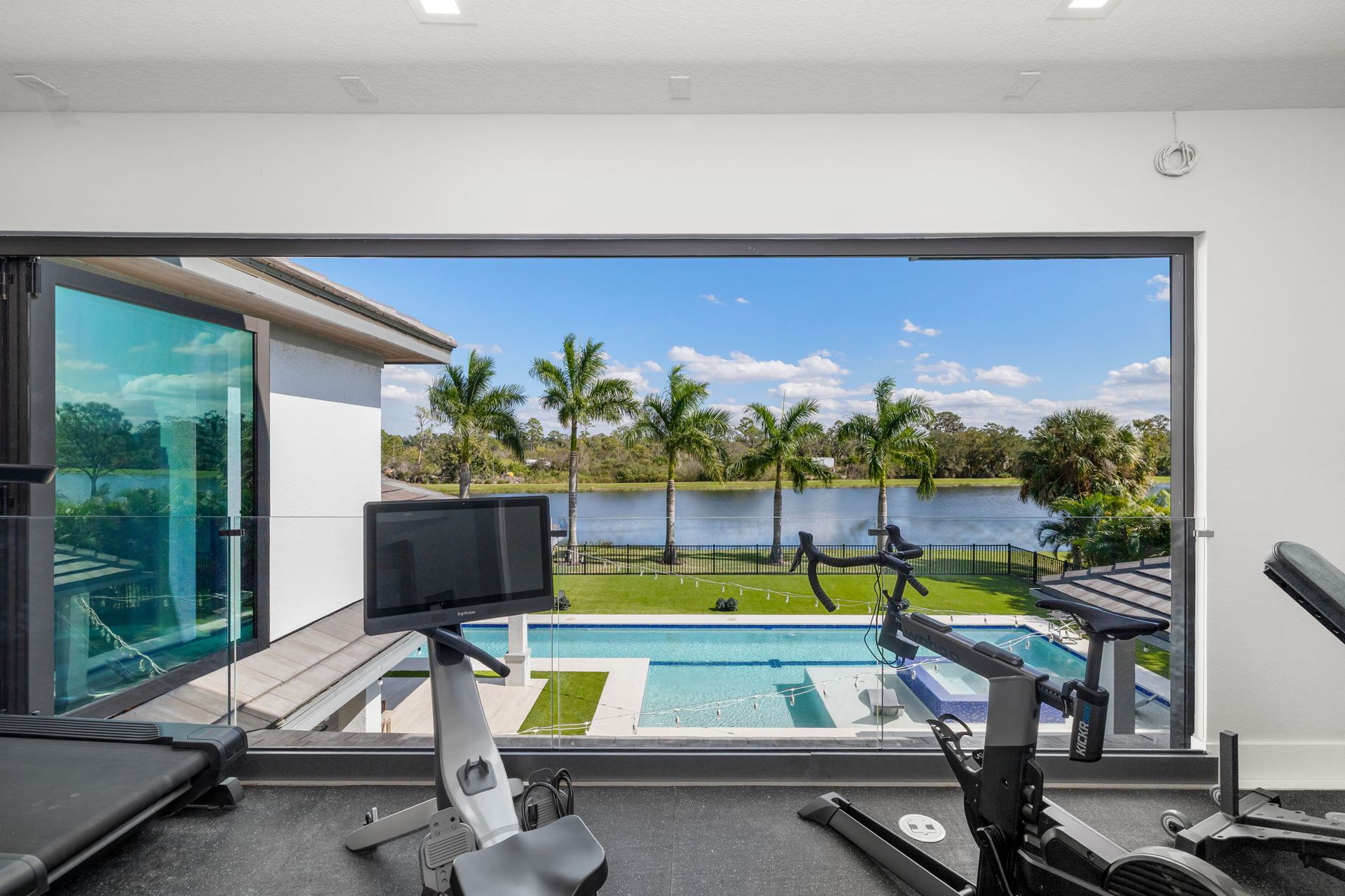 Indoor gym with cardio equipment overlooking a pool, palm trees, and a lake through open glass doors