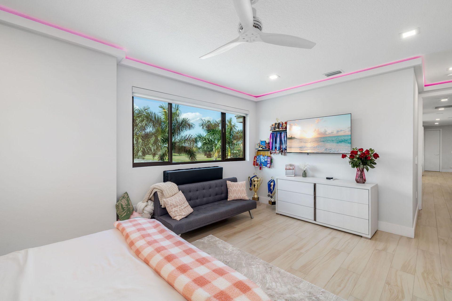 Bright modern bedroom with a bed, gray sofa, dresser, large window, and pink LED ceiling lights