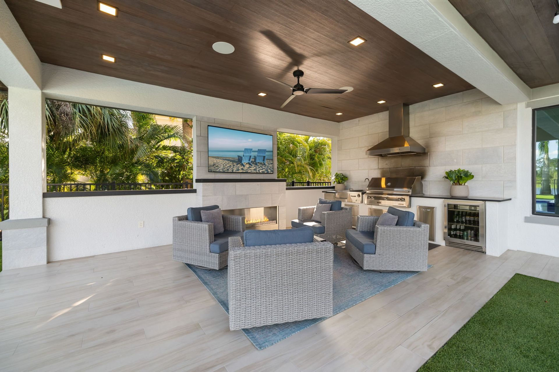 Covered outdoor patio with gray seating, fire pit, grill, and large windows overlooking greenery and water.