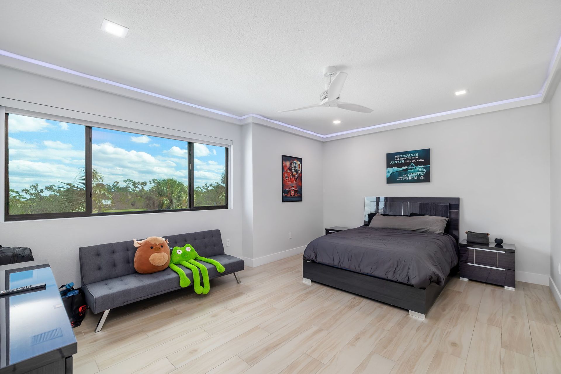 Bright bedroom with gray bed, couch, wall art, large window, and a green toy on the floor