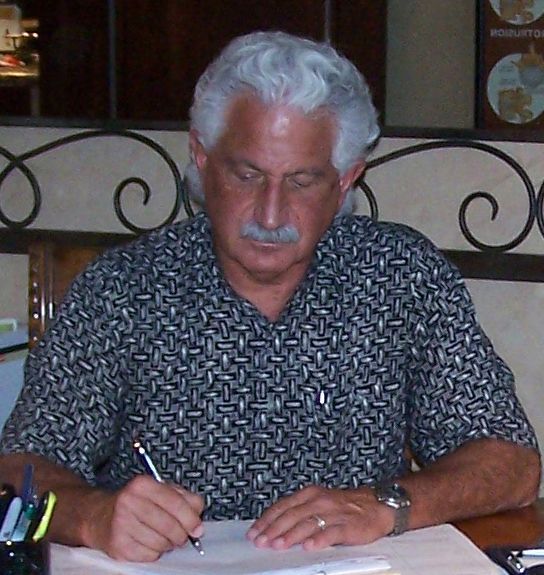 Man with white hair, mustache, writing at desk. Dark shirt, ornate background.