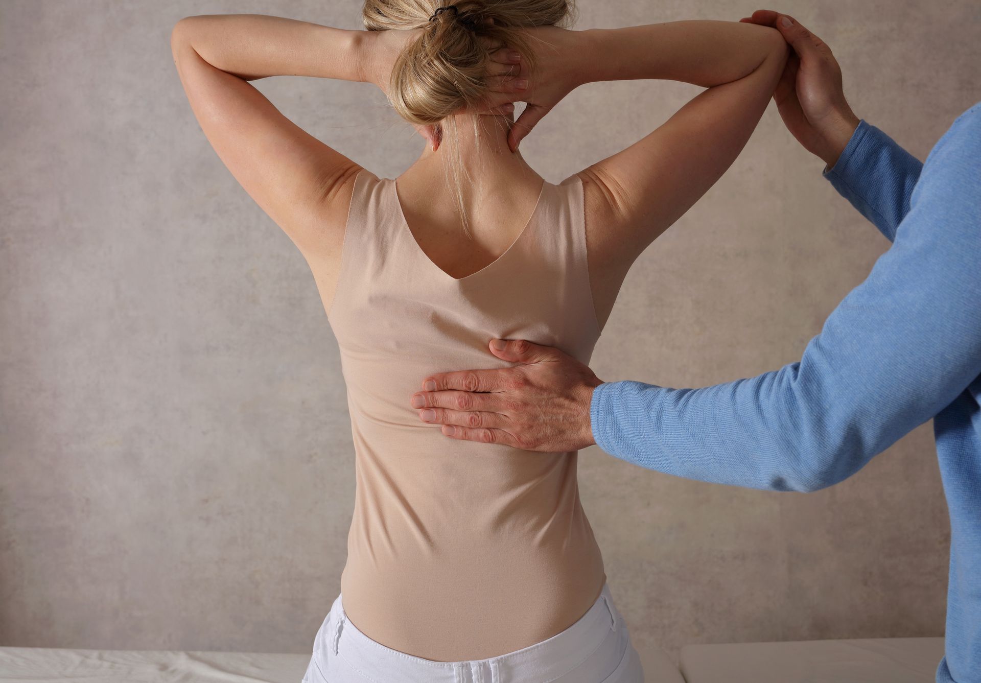 A person's back being examined by a healthcare provider. The person is wearing a beige top and white pants.