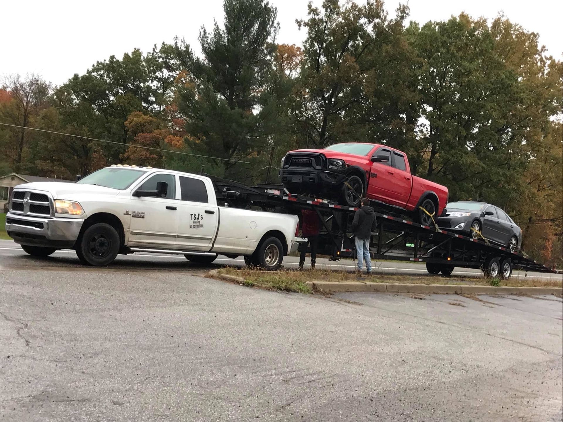 White tow truck hauling red and black pickup trucks on a trailer by a roadside with trees behind.