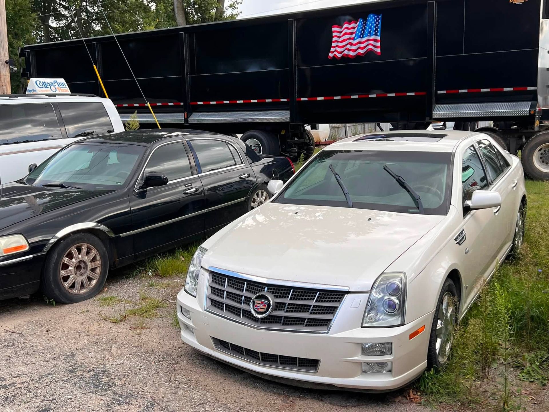 Two parked sedans, black and white, in a gravel lot beside a black trailer with an American flag graphic.