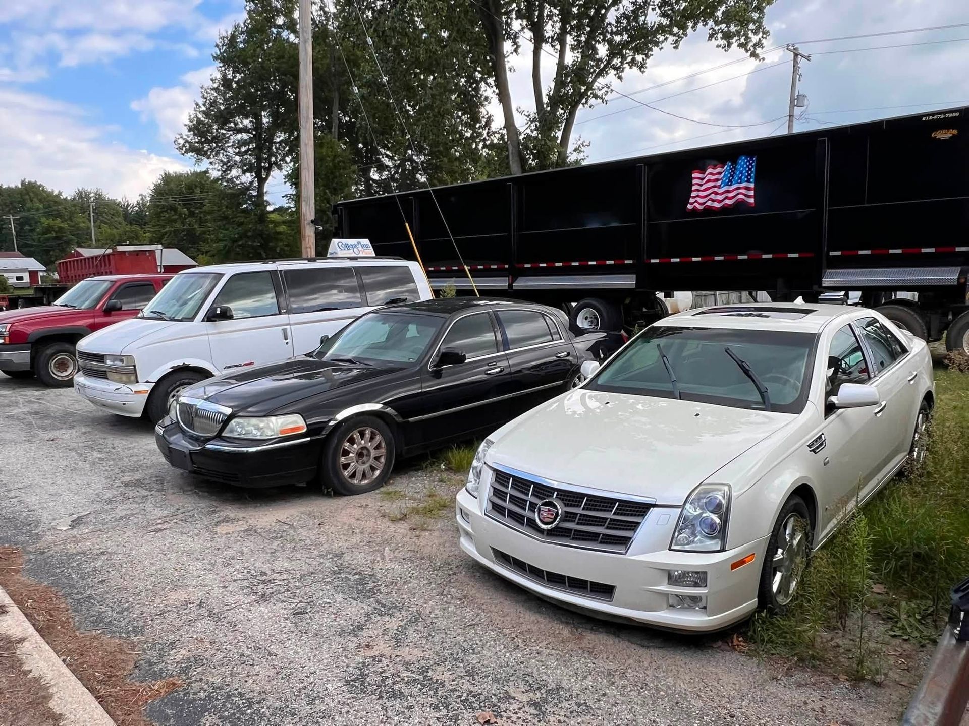 Three parked cars beside a black trailer in a gravel lot, including a white Cadillac in front.