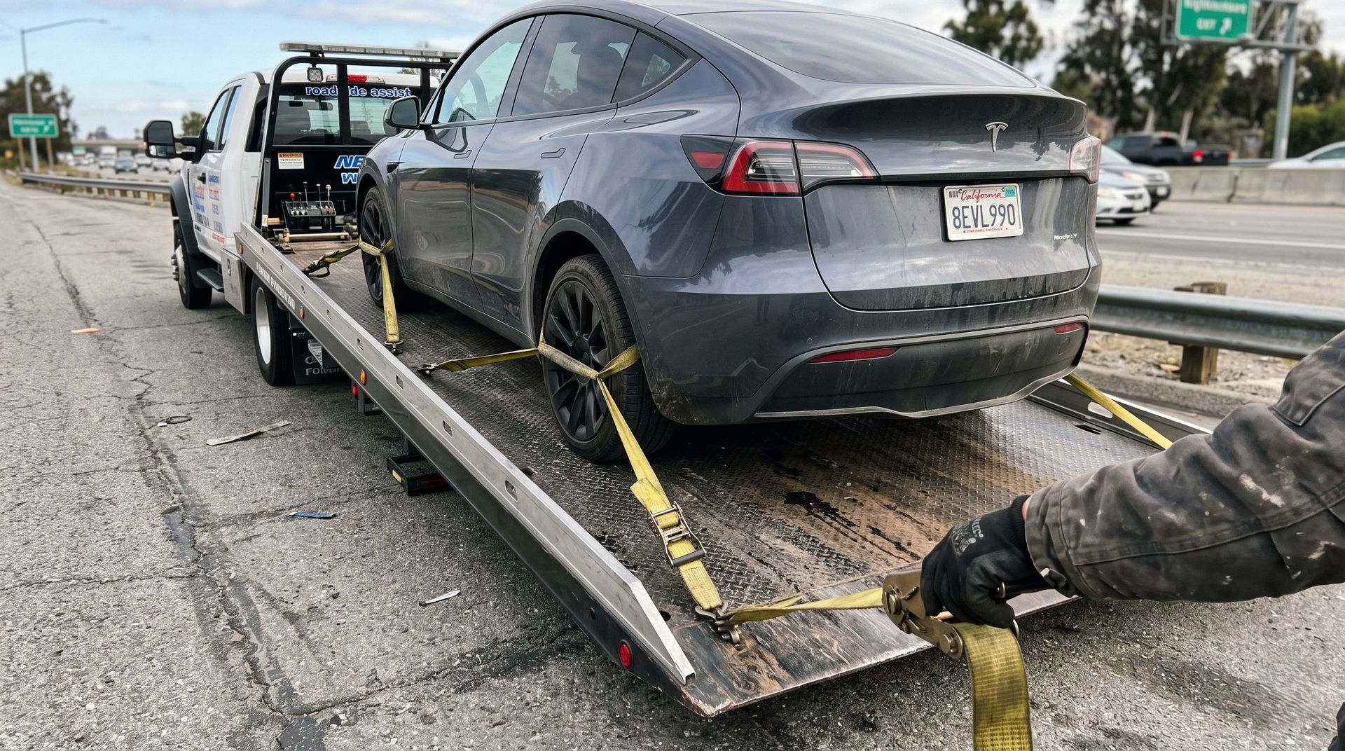 Gray Tesla on tow truck flatbed on roadside, secured with straps after a crash, with a damaged front wheel visible
