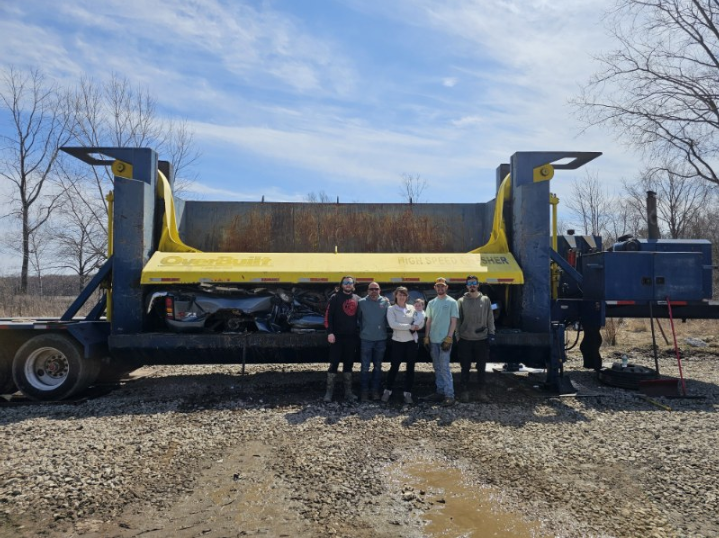 Workers pose beside a large yellow industrial machine on a trailer in a gravel lot.