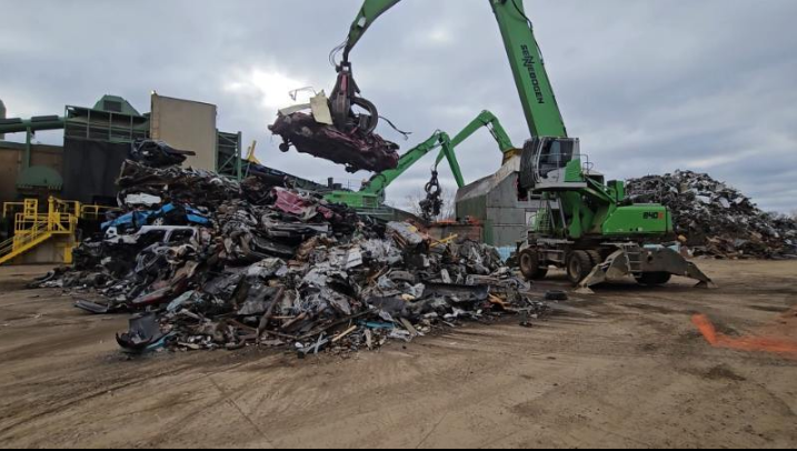 Industrial yard with a green crane lifting scrap metal in a large junk pile under cloudy skies