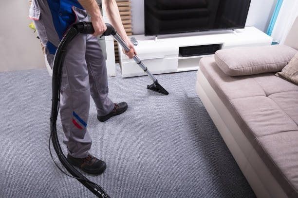 Person vacuuming a gray carpet beside a beige sofa in a living room