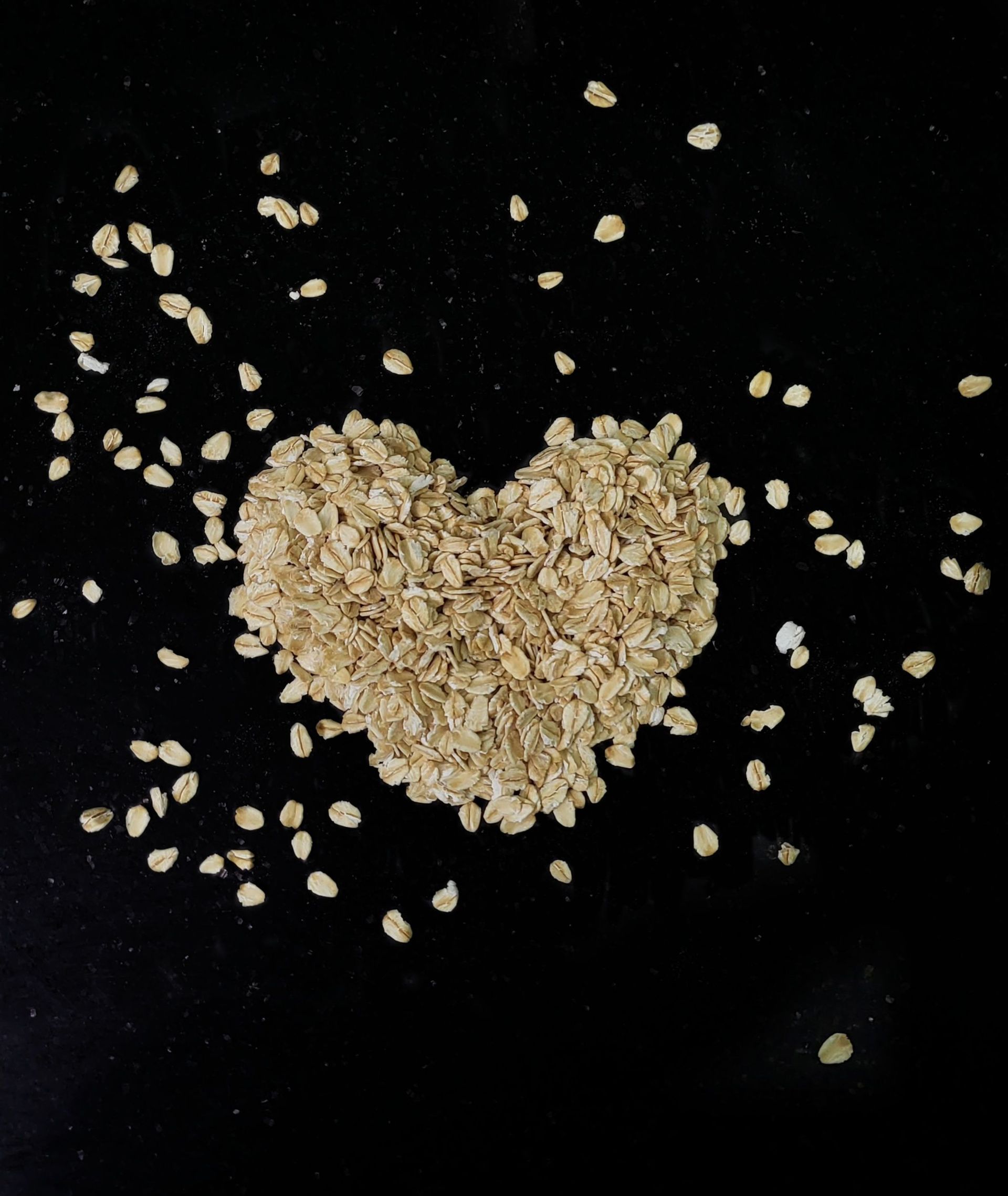 Heart-shaped pile of oatmeal flakes on a black background, with scattered oats around it