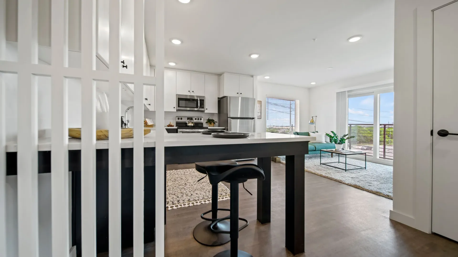 Open-concept kitchen with white cabinets, a dark island, and a living area with large windows.