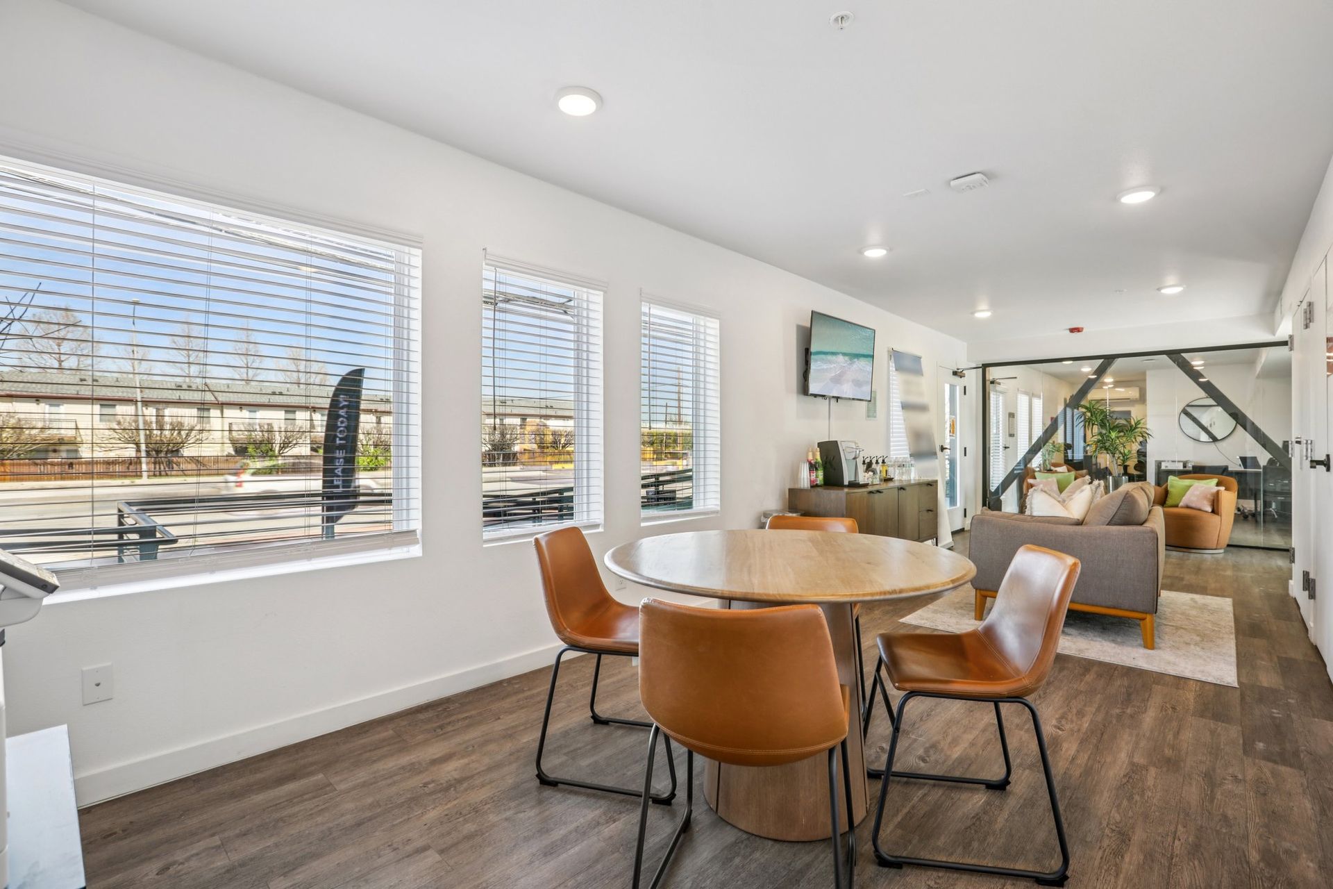 A modern, brightly lit office or lounge area with a round wooden table, four tan chairs, and nearby seating furniture.