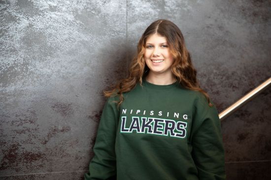 Man wearing a Lakers sweatshirt and beanie leaning against a wall in a hallway.