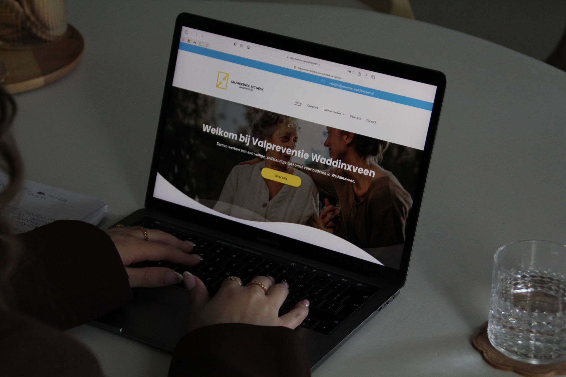 Een persoon typt op een laptop met een website open, naast een glas water op een bureau.