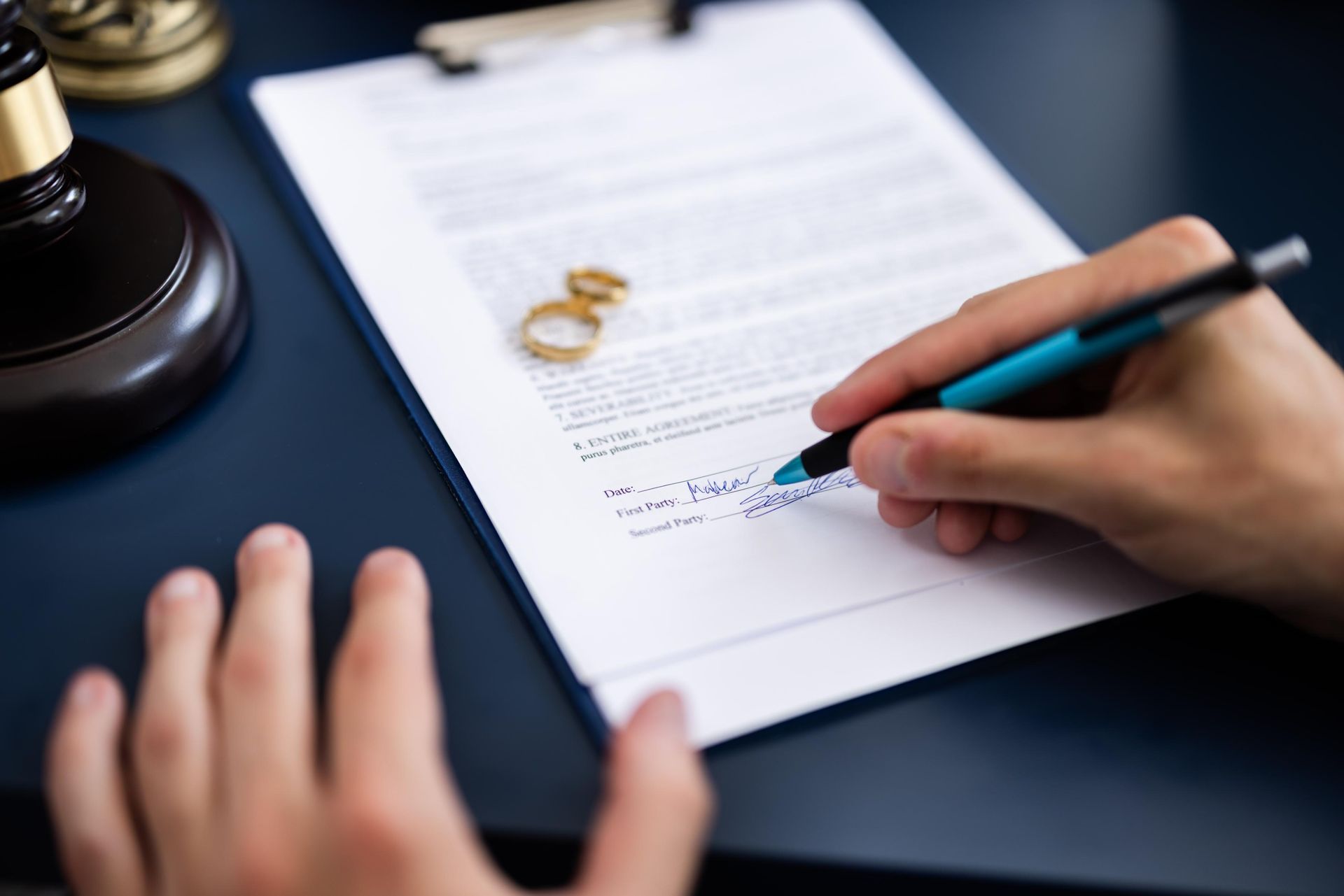 Person signing a document, with wedding rings and gavel in the background.