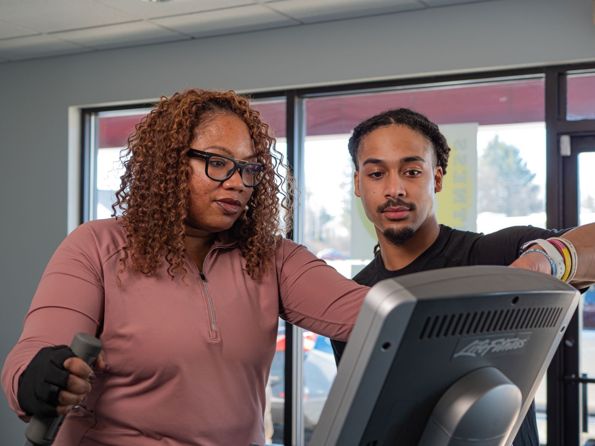 Woman in glasses on elliptical machine with trainer in gym, looking at the machine's display.