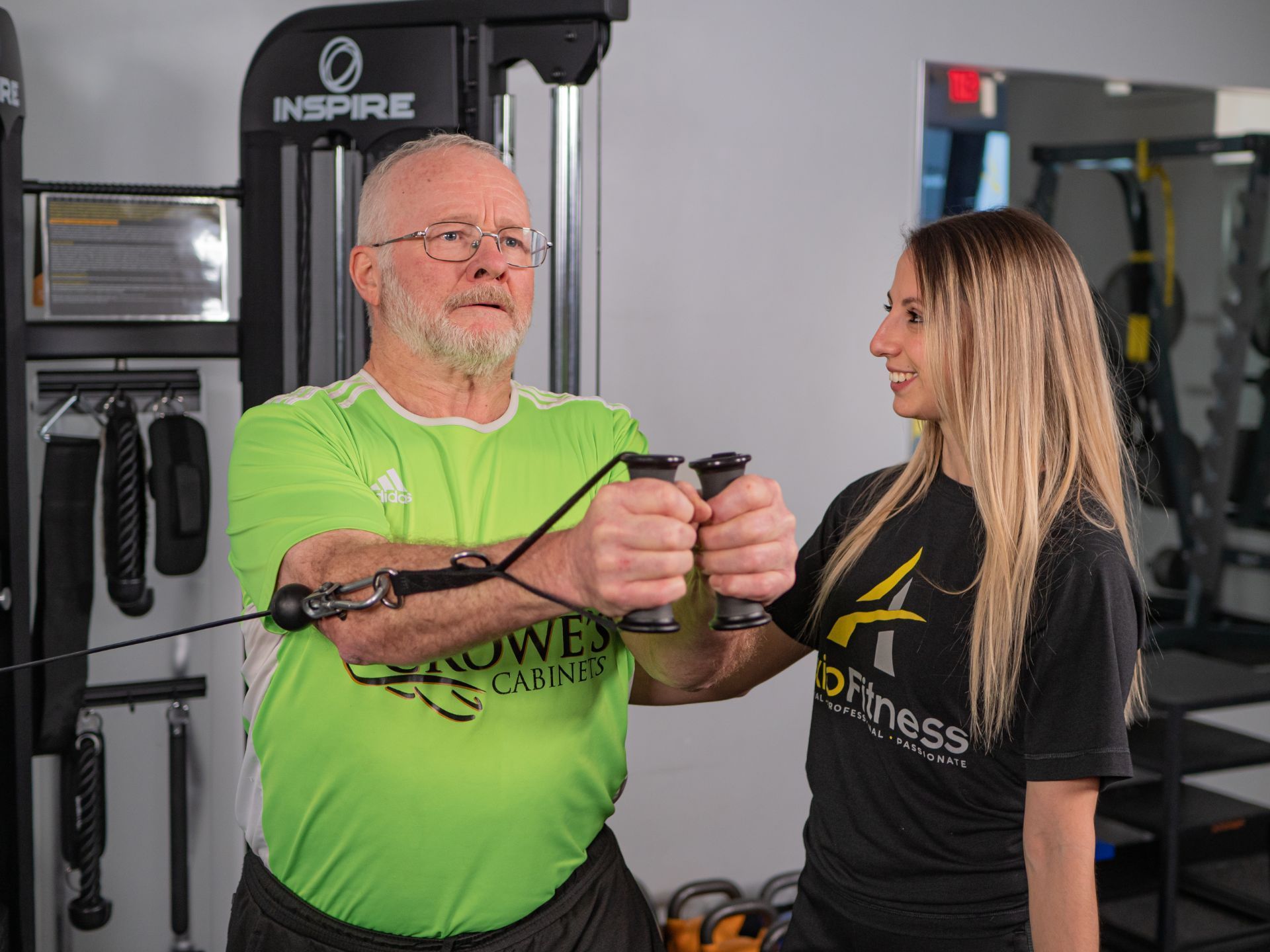 An older man in a green shirt works out at a gym with a personal trainer. The trainer helps him with cable chest flys.