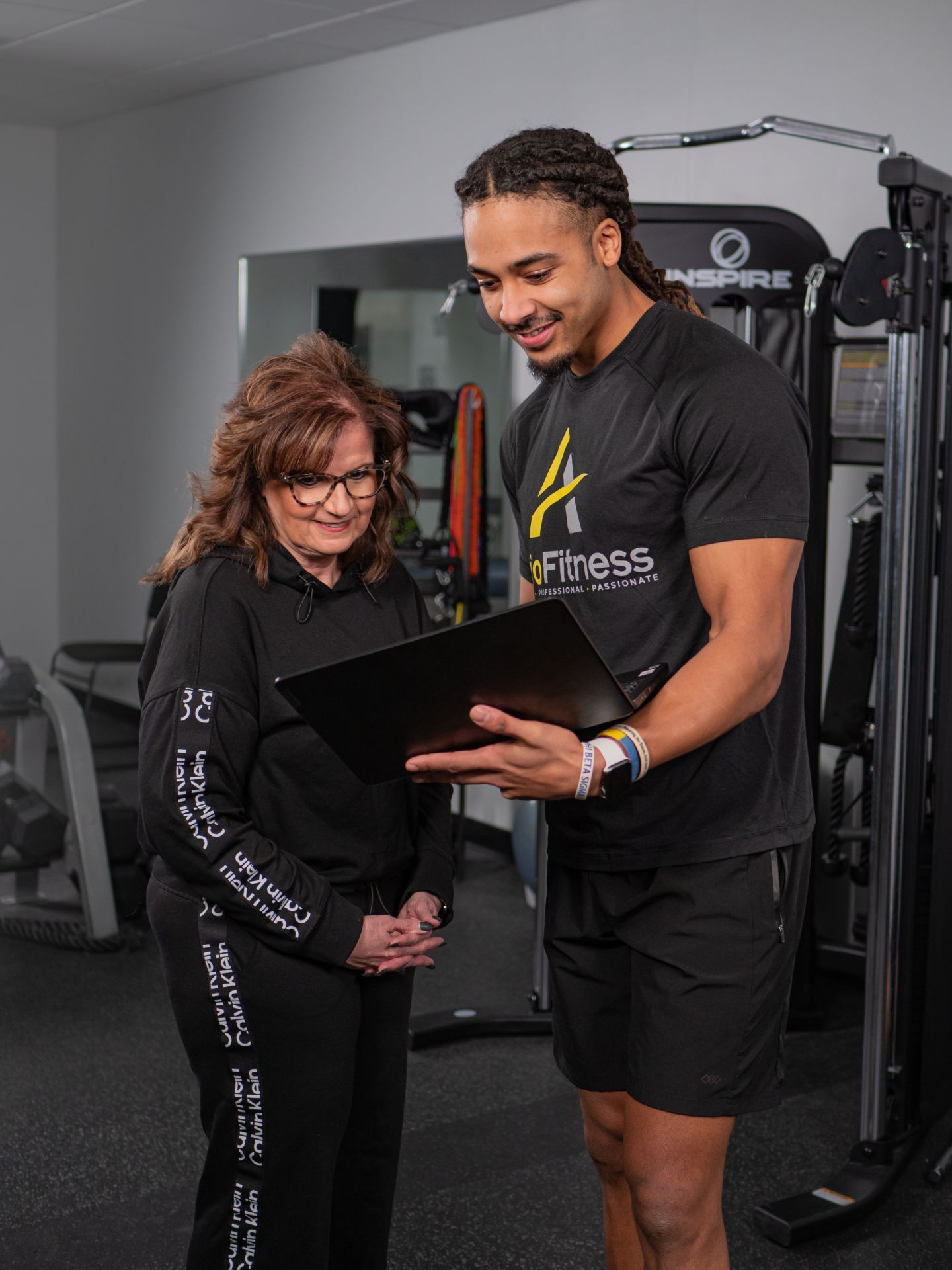 A young fitness trainer shows a digital tablet to an older woman in a gym. Both are smiling.