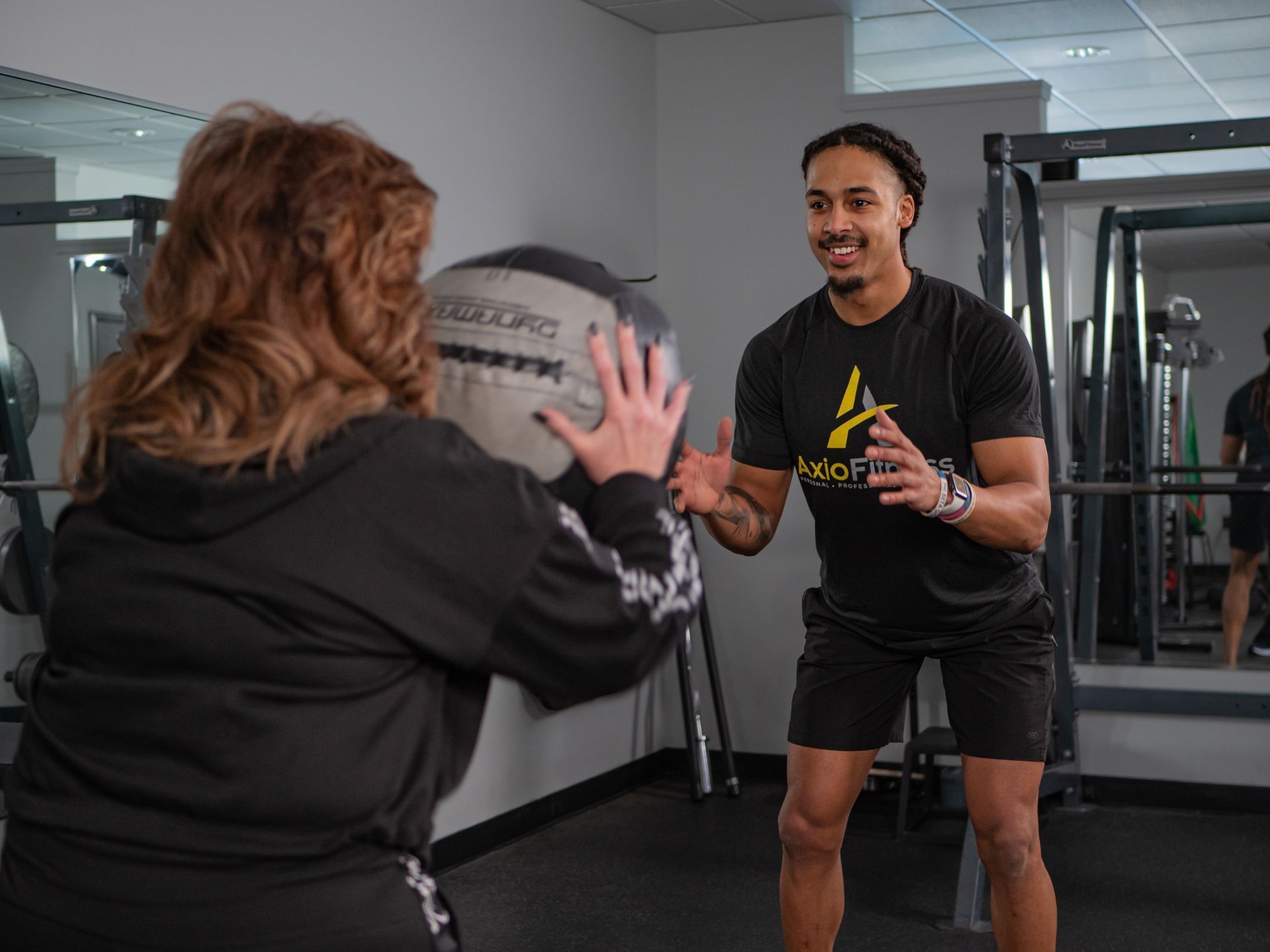 A trainer and a woman are in a gym. The trainer, smiling, is about to catch a medicine ball from the woman.