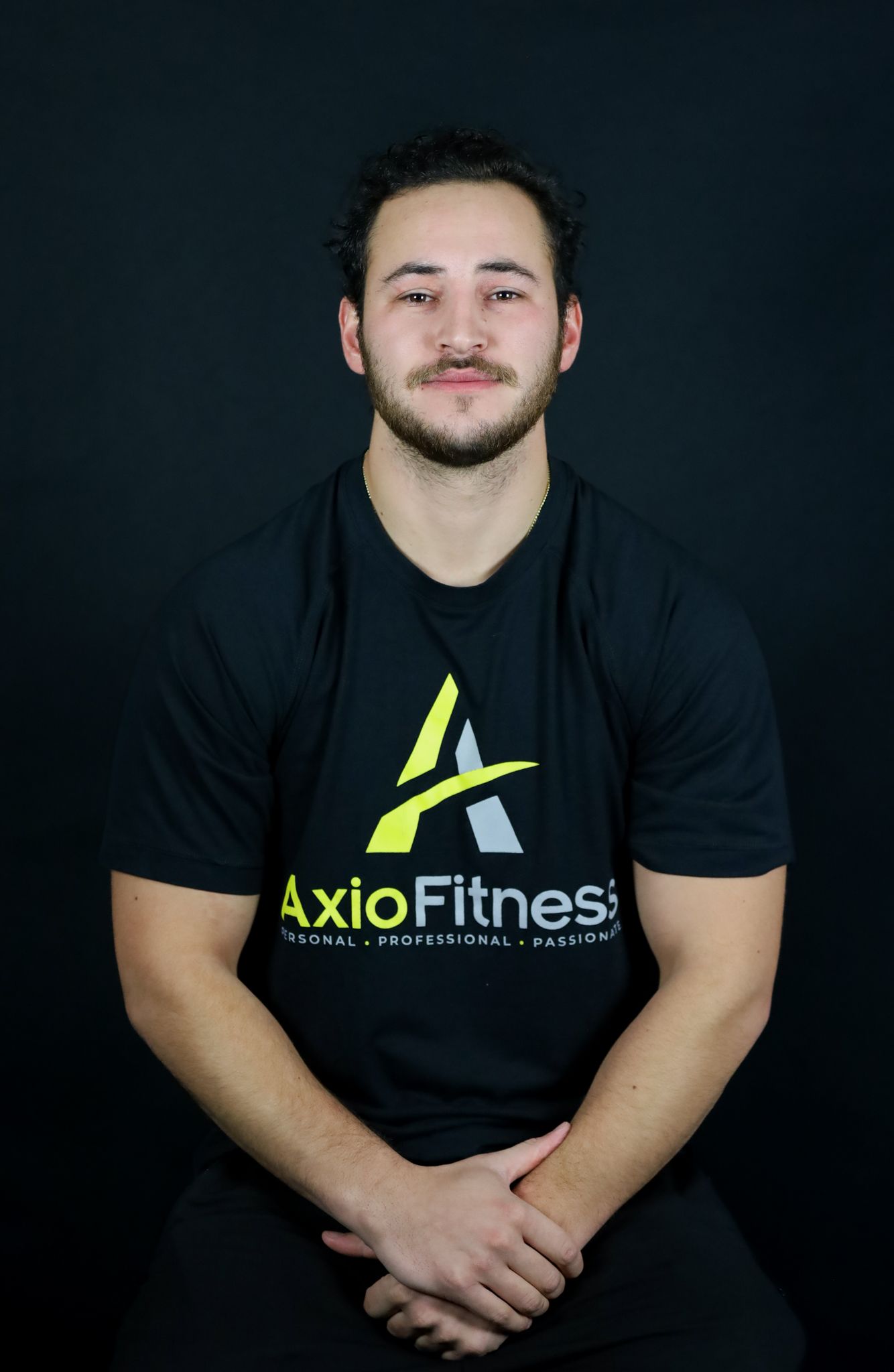 Man in a black AxioFitness t-shirt, seated and smiling, against a black backdrop.