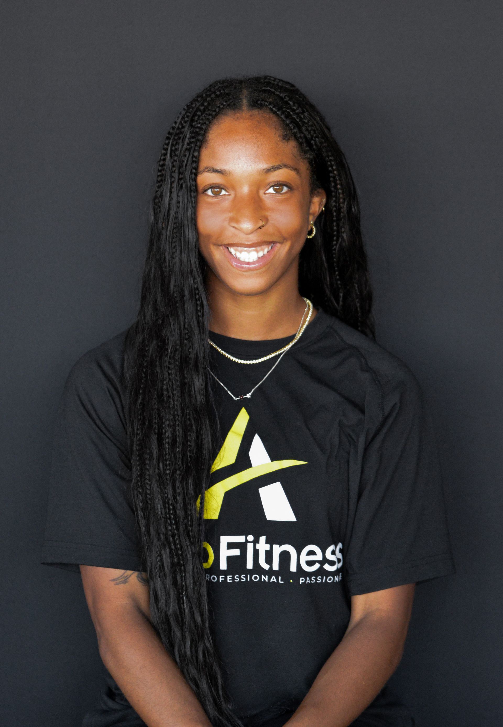 A young Black woman with long braids, wearing a black t-shirt with a logo, smiles at the camera against a dark background.