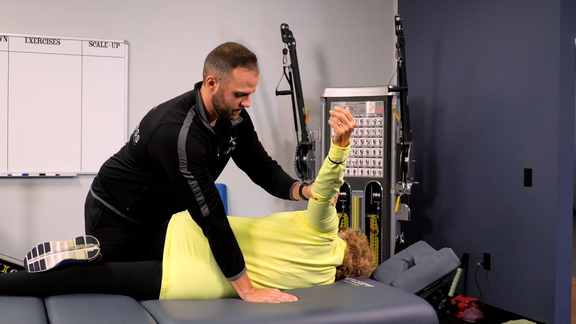 A man assists a woman lying on a table with arm raised during a physical therapy session in a clinic.