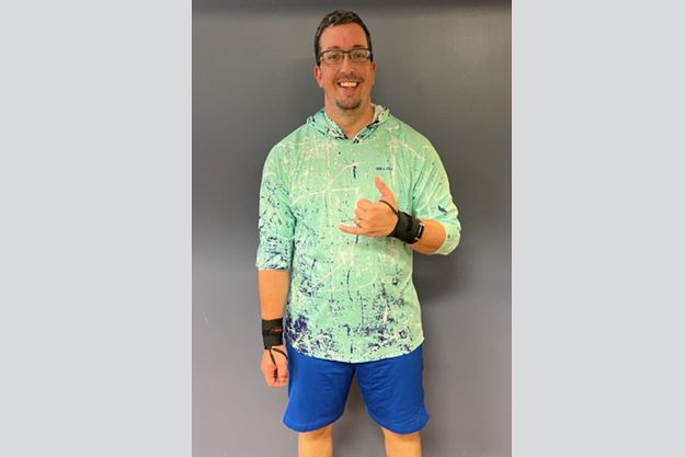 Man wearing a green and blue patterned hoodie, blue shorts, and wrist supports, giving a shaka sign. 