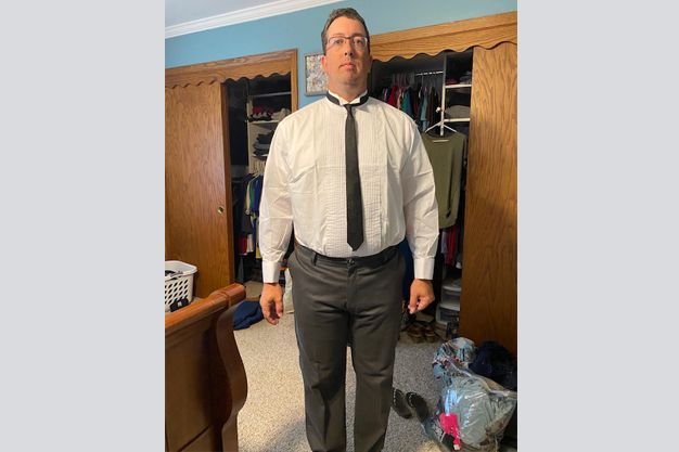 Man in white shirt and black tie, standing in a room with two open closets. He is wearing black pants and glasses.
