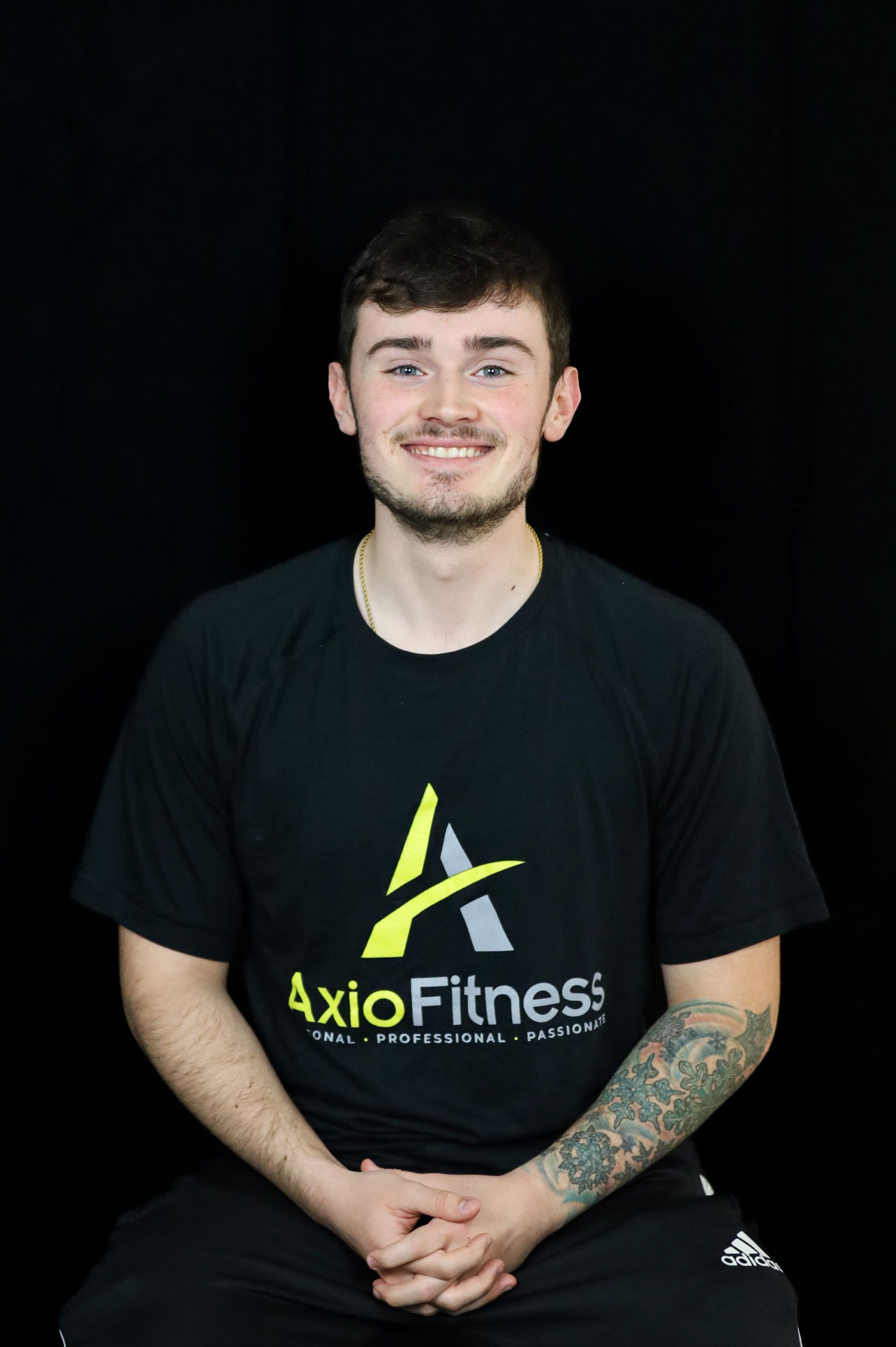 Man with arm tattoos wearing a black AxioFitness t-shirt, smiling, seated in front of a black backdrop.