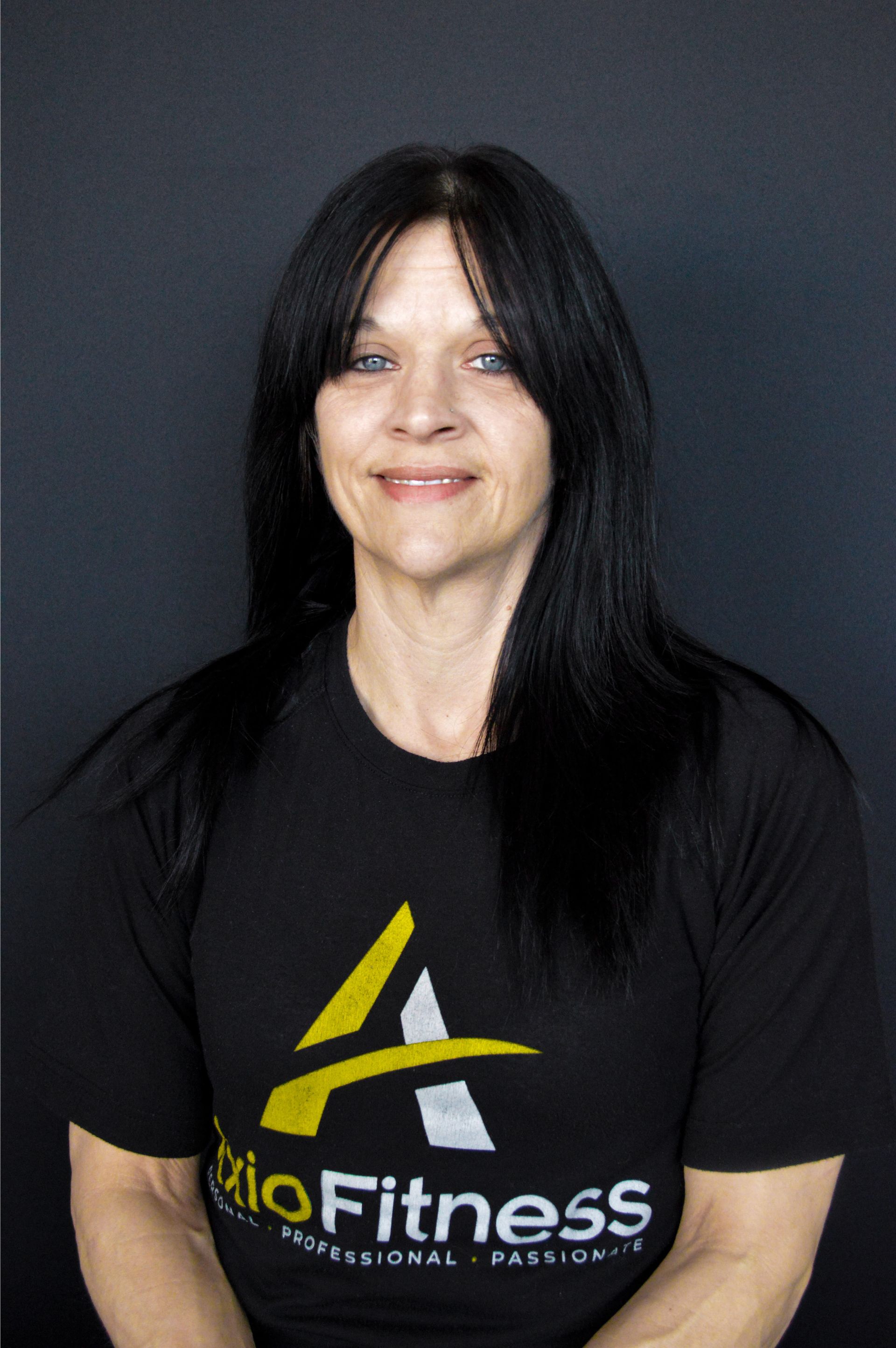 Woman with black hair wearing a black t-shirt with a yellow logo, smiles at the camera against a dark background.