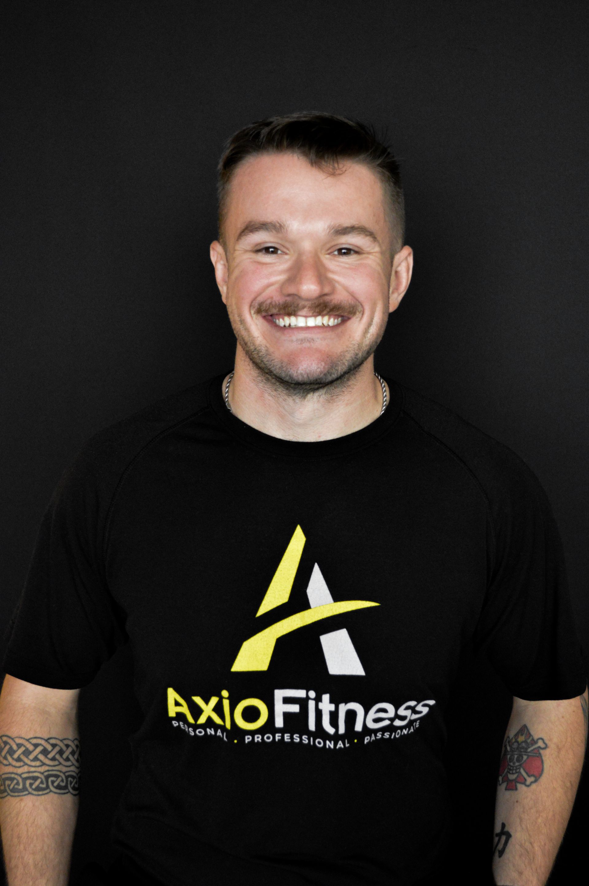 A smiling man with a mustache, wearing a black t-shirt with a yellow and white logo, standing in front of a black background.