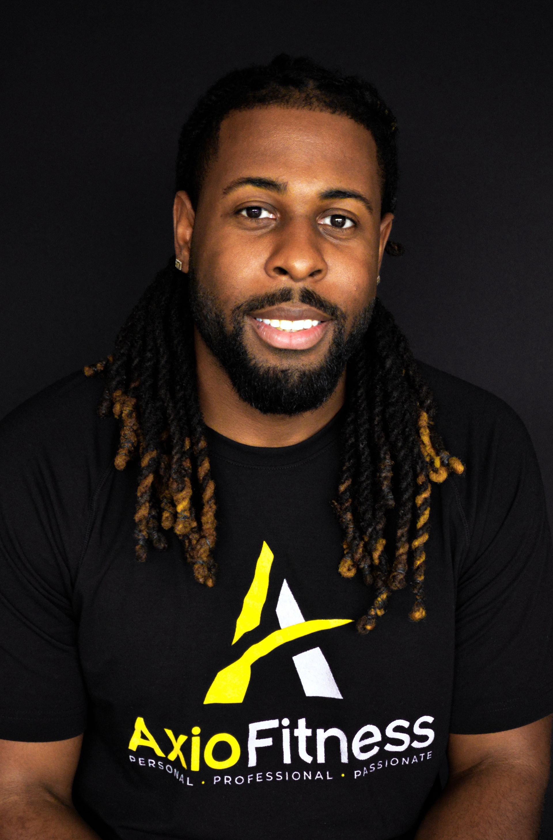 A Black man with long, brown dreadlocks and a beard smiles at the camera. He wears a black Axio Fitness t-shirt.