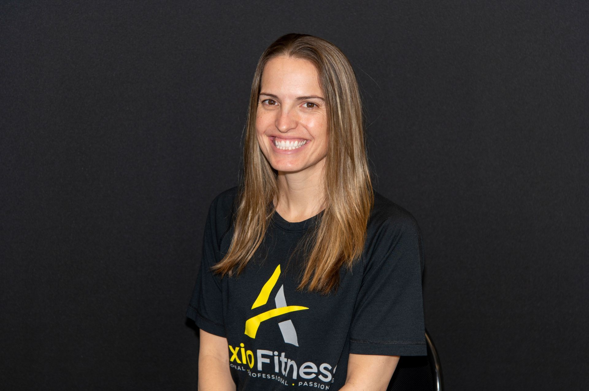 Woman with long brown hair smiling, wearing a black t-shirt with a yellow logo, sitting against a black backdrop.