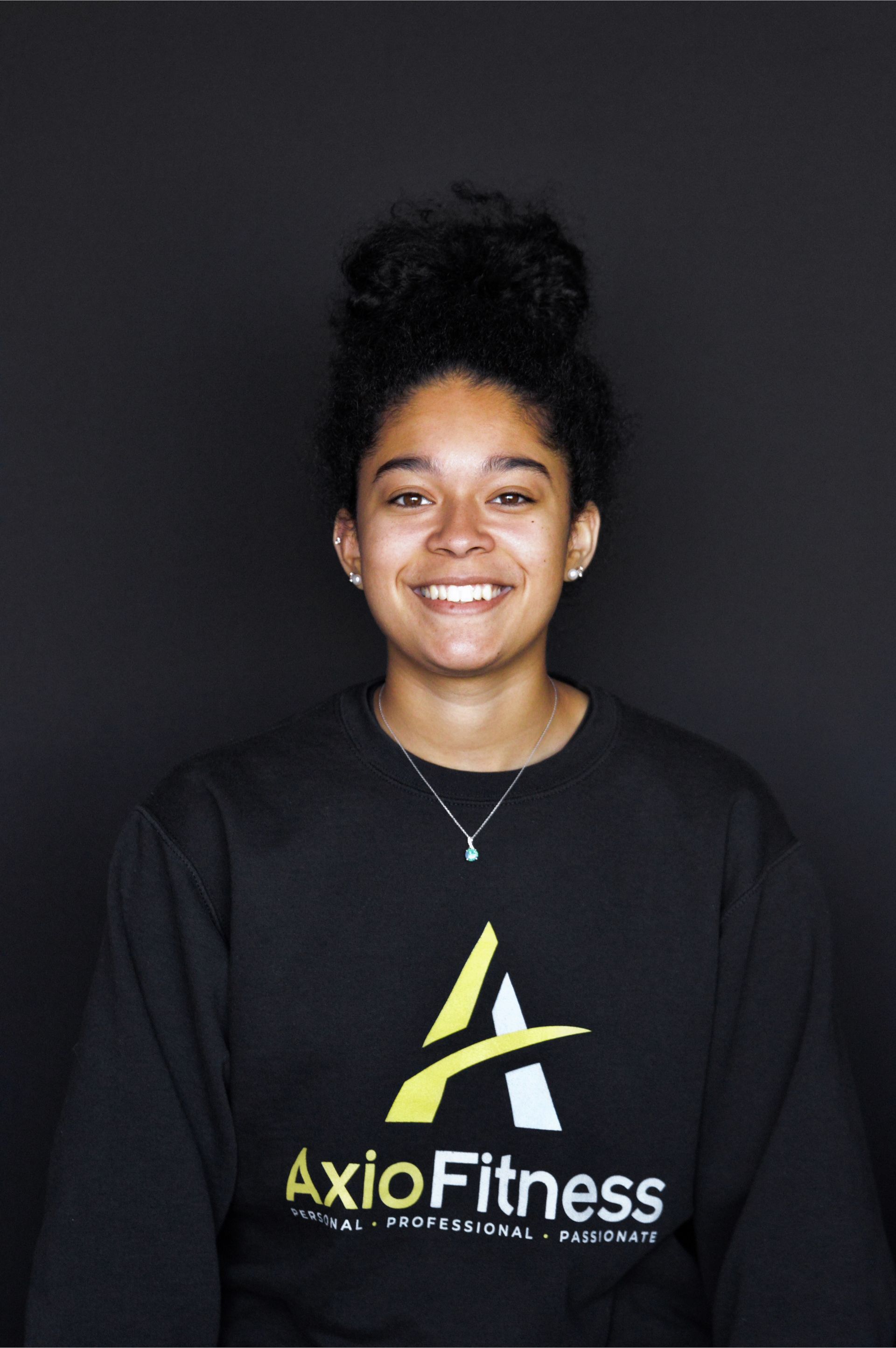 Woman wearing a black AxioFitness sweatshirt, smiling, hair in a bun, on a black background.