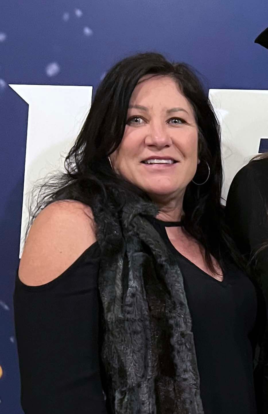 Woman with dark hair, wearing a black off-the-shoulder top and a grey scarf, smiles at the camera.