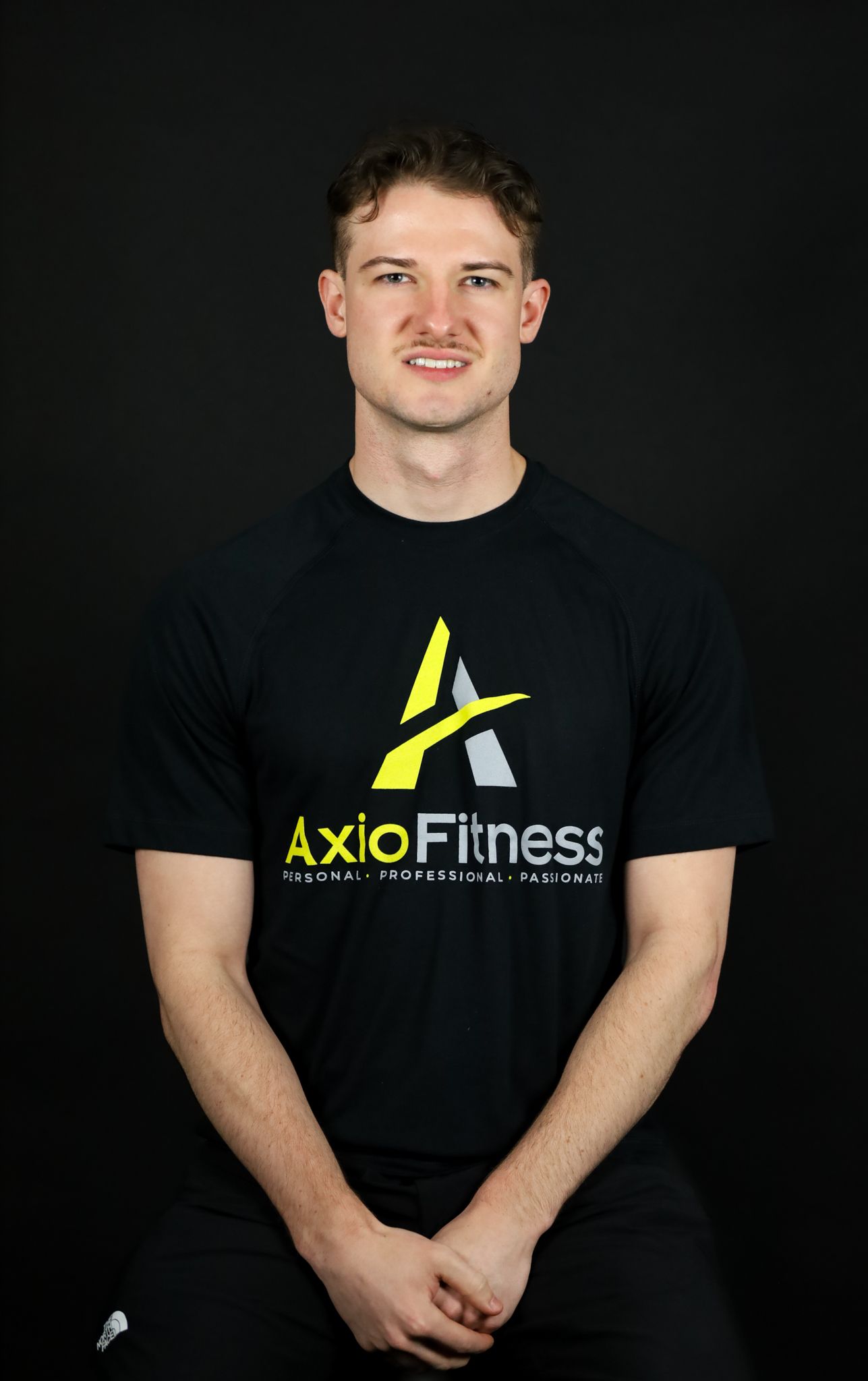Man in a black AxioFitness t-shirt, smiling, sitting against a black background. Arms are crossed.