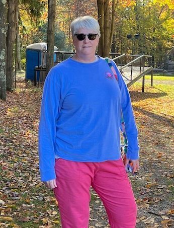 Woman with short gray hair wearing sunglasses, a blue long-sleeved shirt, and pink pants.