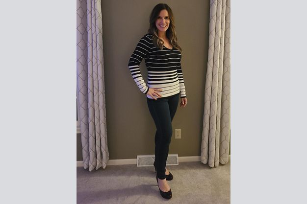 Woman with brown hair, wearing a striped top, dark jeans, and heels, poses smiling in front of curtains.