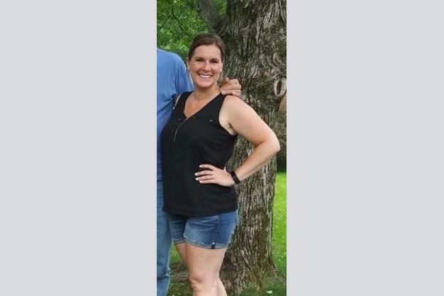 Woman in black top and jean shorts poses with a hand on her hip, smiling outdoors near a tree.