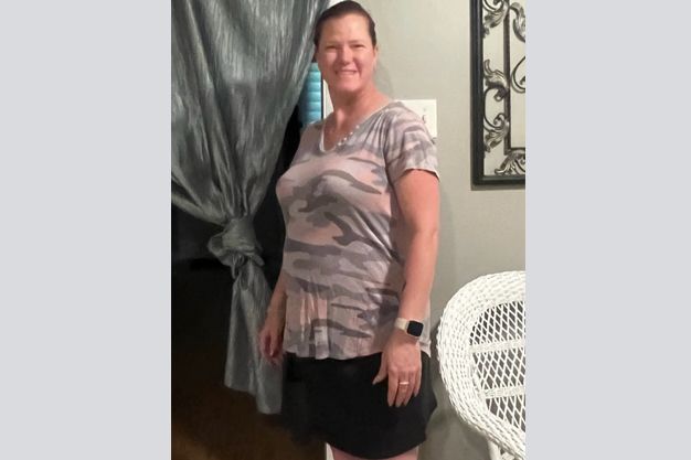 Woman in camouflage top and black skirt, smiling, standing indoors near a curtain and a white chair.