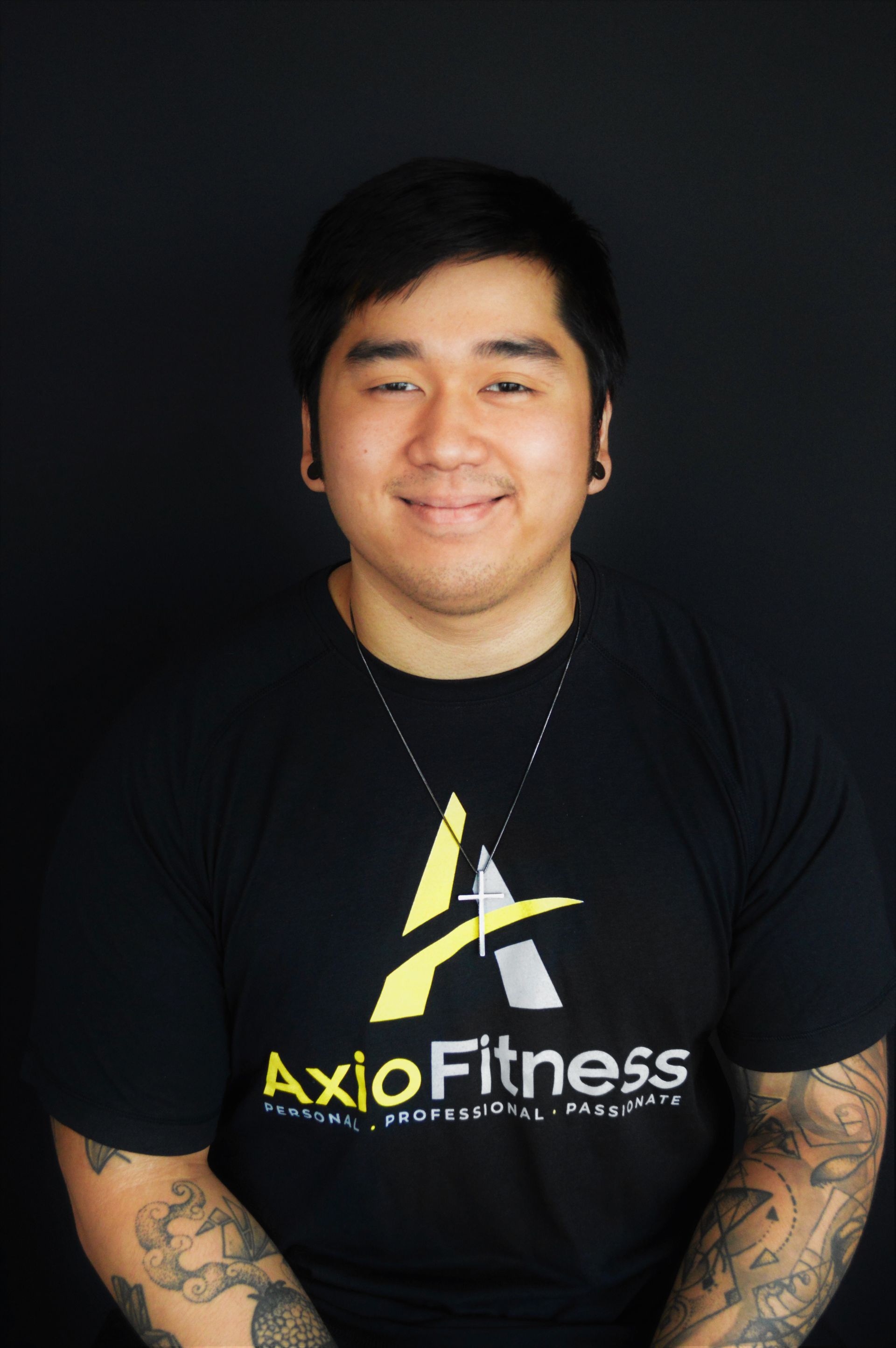 Smiling Asian man in a black AxloFitness shirt with tattoos on his arms, posing against a black backdrop.