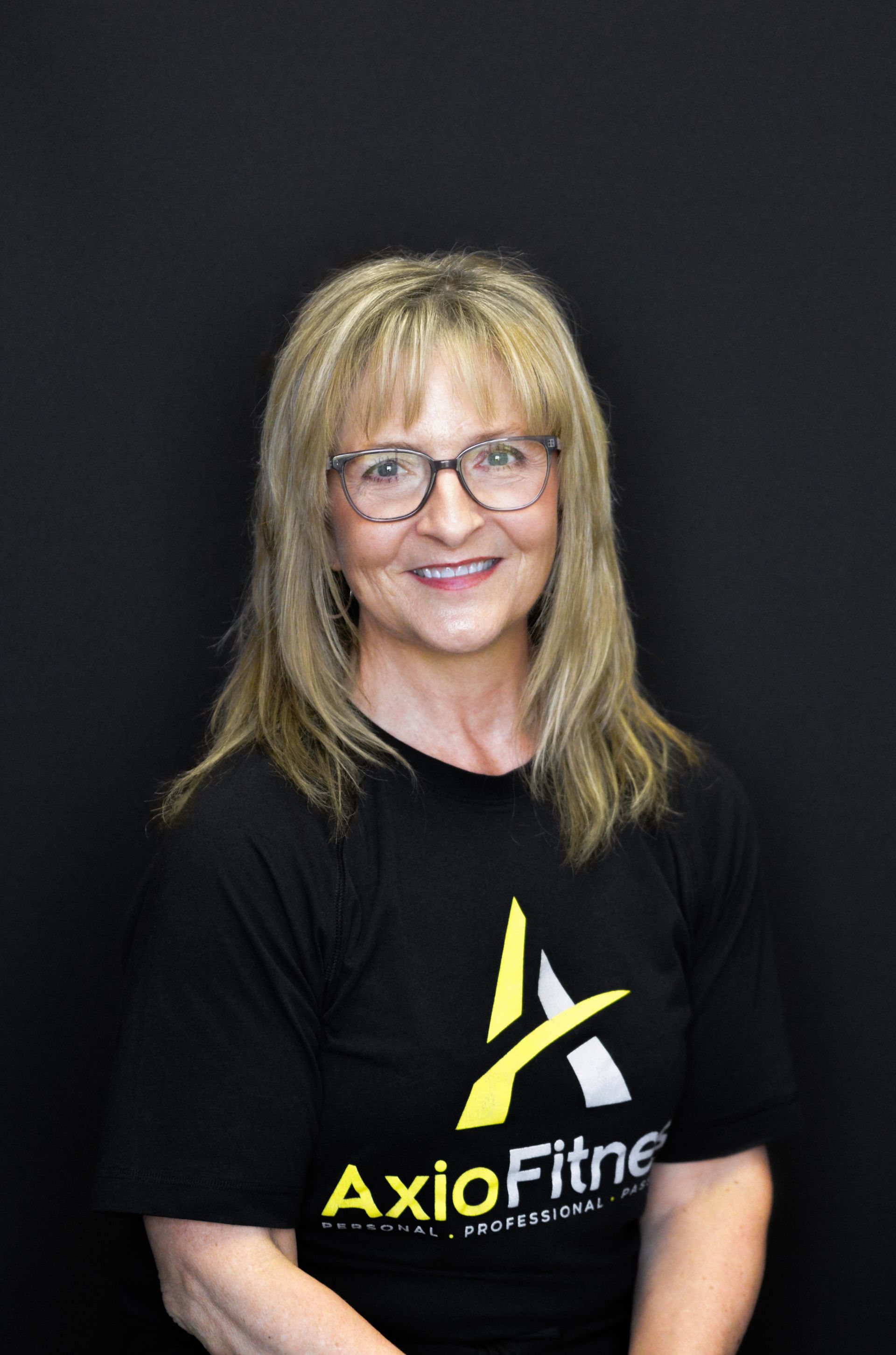 Woman with blonde hair and glasses wearing a black Axio Fitness shirt, smiling at the camera against a black backdrop.