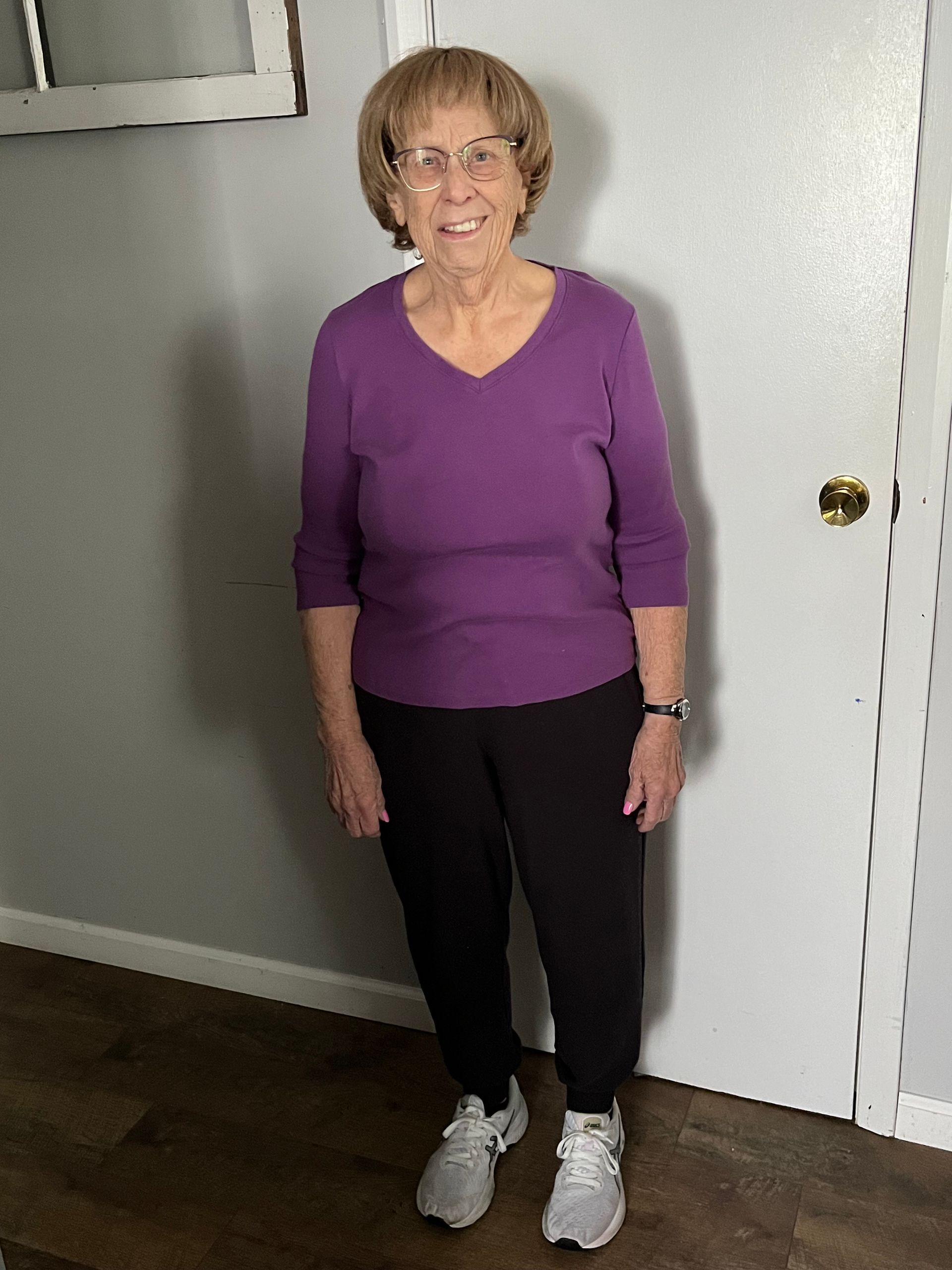 An elderly woman with short brown hair and glasses stands indoors, wearing a purple top, black pants, and sneakers.