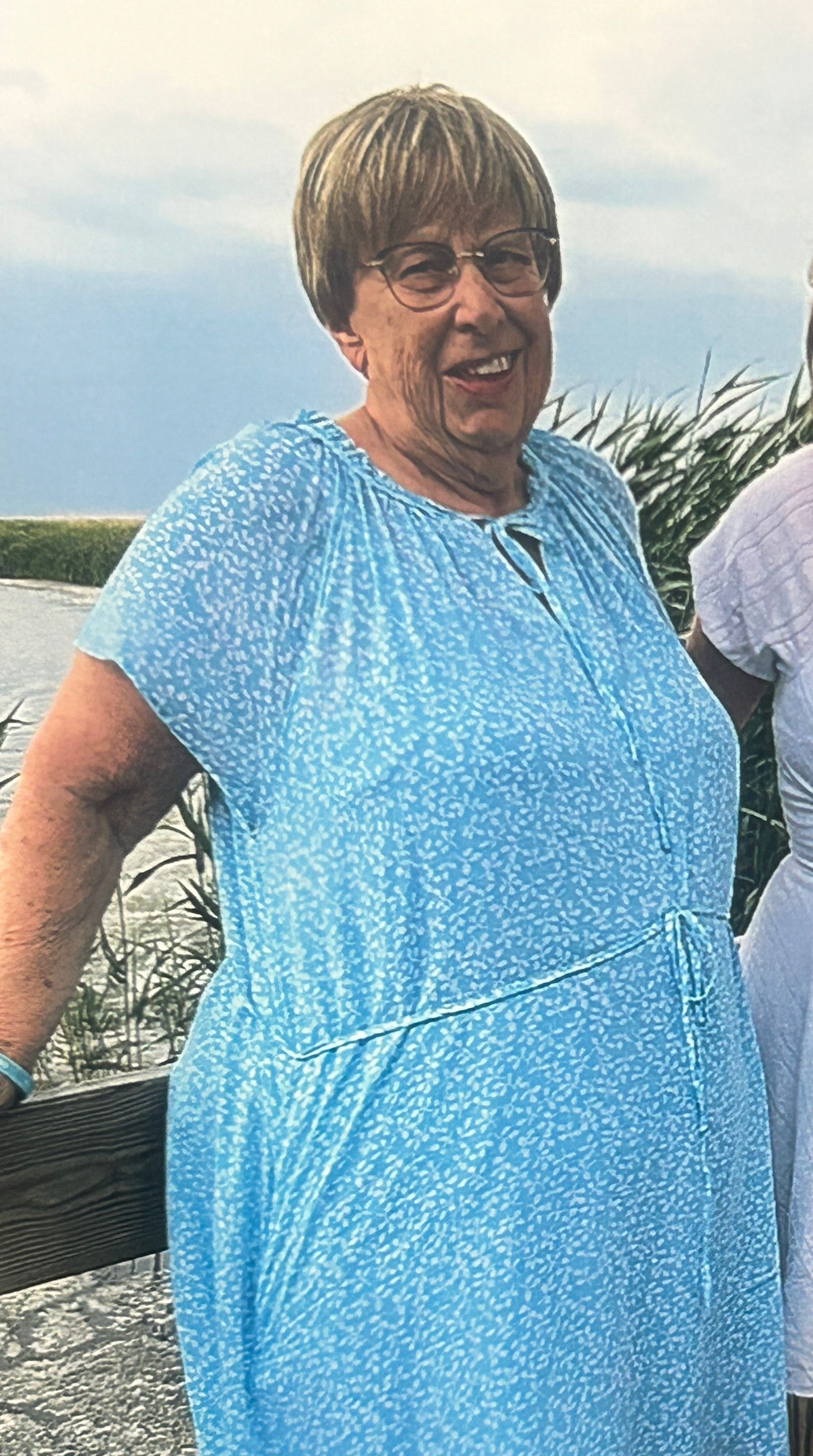 Elderly woman in a blue floral dress, smiling, standing by a body of water. 