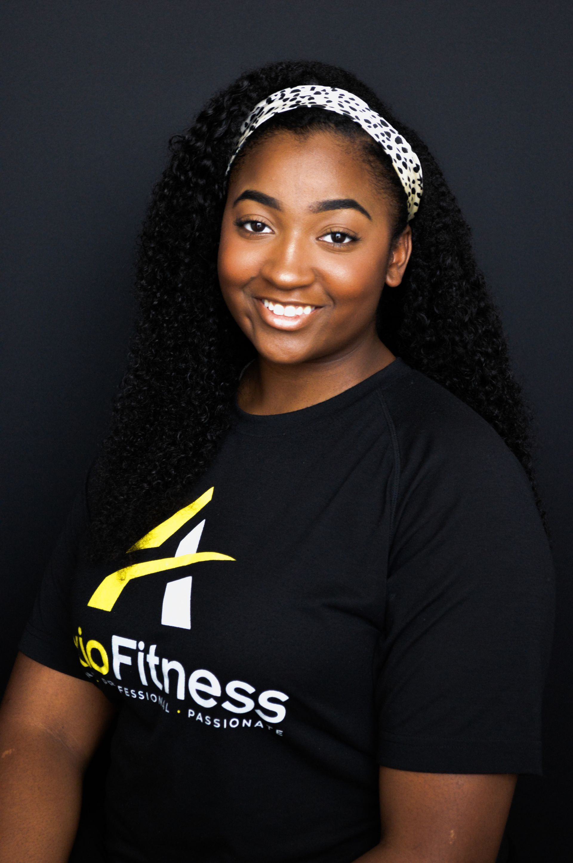 A smiling Black woman with long curly hair wearing a headband and a black t-shirt with a logo.