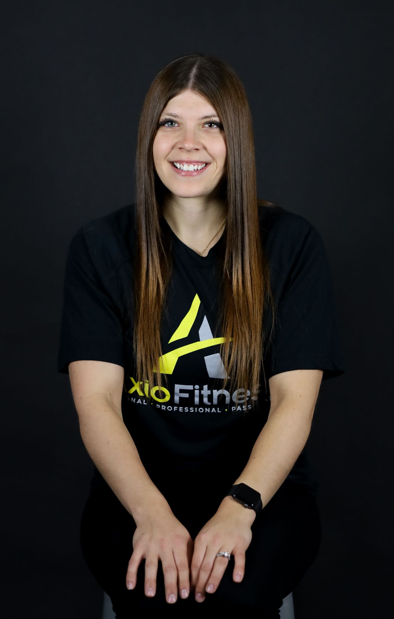 A Woman wearing a black t-shirt with a yellow and black logo, and a black watch, seated against a dark background.