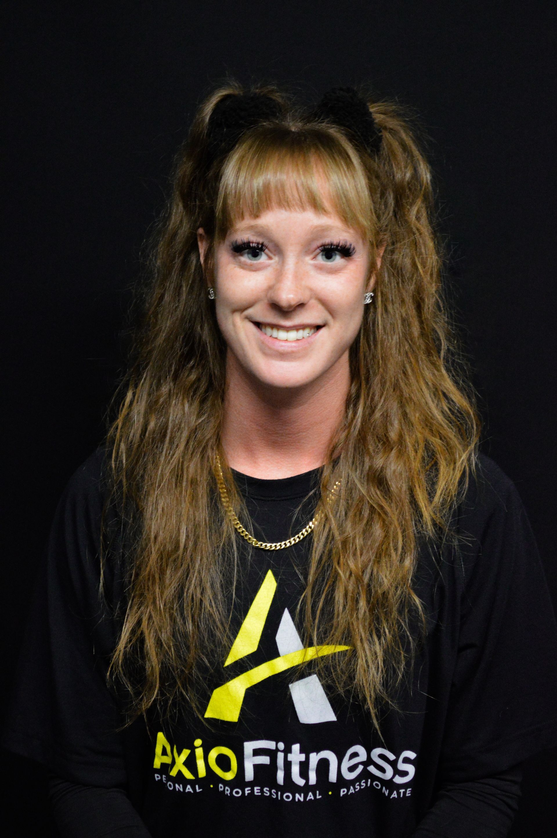 A smiling person with long light brown hair wears a black AxioFitness t-shirt against a black background.